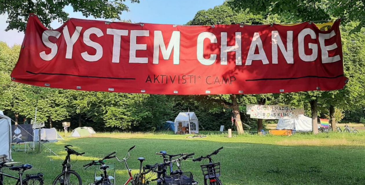 Foto: Im Vordergrund ein Banner mit systemchange not climate change, darunter Fahrräder. Im Hintergrund eine Wiese und Zelte